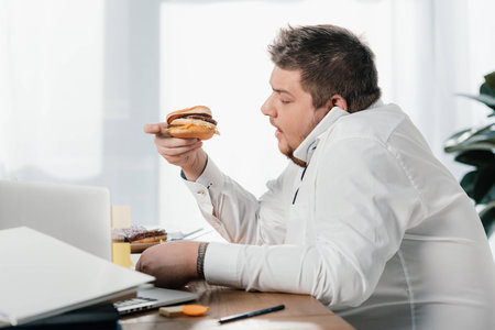 overweight businessman eating hamburger while working in officeの写真素材