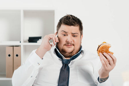 overweight businessman talking on smartphone while eating hamburger in officeの写真素材