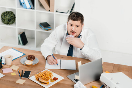 overweight businessman eating donuts, hamburger and french fries while wokring in officeの写真素材