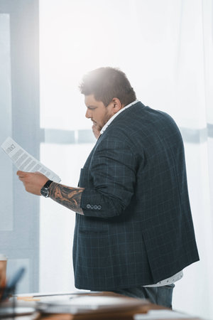 overweight businessman in suit looking at document while standing at window in officeの写真素材