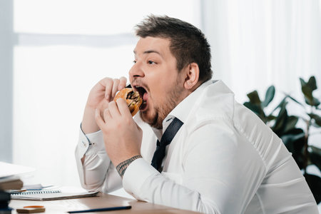 fat businessman talking on smartphone while eating hamburger in office