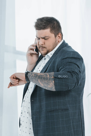 overweight businessman looking at watch while talking on smartphoneの写真素材