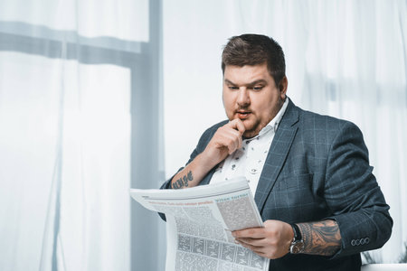 thoughtful overweight businessman in suit reading newspaper in officeの写真素材