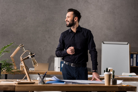 smiling architect standing near working table and looking away