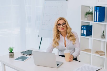 beautiful businesswoman sitting at table with laptop and holding disposable coffee cupの写真素材