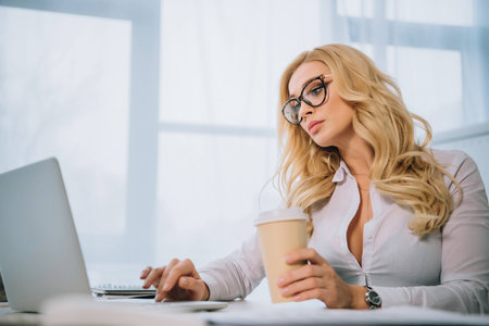 beautiful businesswoman working at laptop in office and holding coffee in paper cupの写真素材