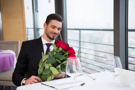 smiling man in suit with bouquet of red roses waiting for girlfriend in restaurantの写真素材