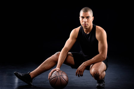 handsome african american basketball player sitting with ball on blackの写真素材