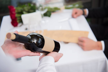 selective focus of waiter showing bottle of wine to visitor at table in restaurantの写真素材