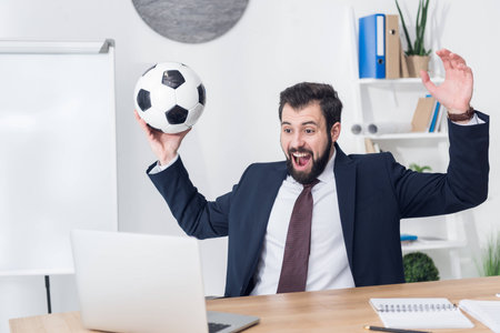 excited businessman in suit with soccer ball looking at laptop screen at workplace in officeの写真素材