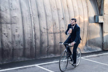 smiling businessman in suit with coffee to go in hand riding bicycle