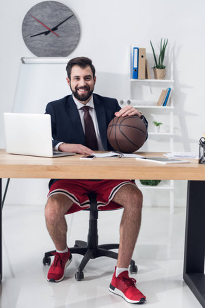 smiling businessman in shorts and jacket with basketball ball sitting at workplaceの写真素材