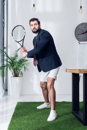 young businessman in jacket and shorts playing tennis in officeの写真素材