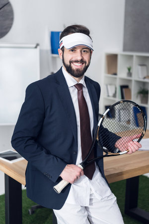 portrait of cheerful businessman with tennis equipment in officeの写真素材