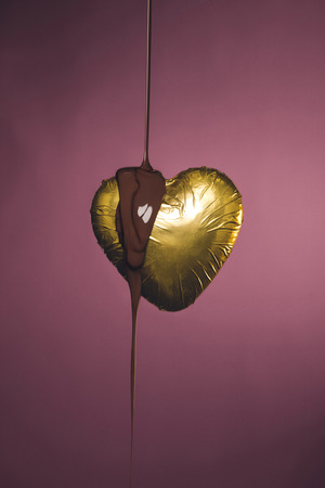 heart shaped candy in golden wrapper with pouring liquid chocolate isolated on pinkの写真素材