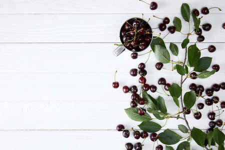 top view of ripe cherries in cup and spilled on white wooden surface with leaves