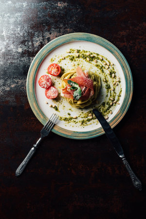 elevated view of pasta with mint leaves, jamon, pine nuts, pesto and cherry tomatoes covered by grated parmesan on plate with fork and knife at tableの写真素材