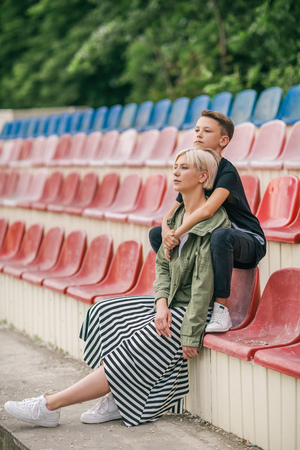 beautiful pensive mother and son hugging and looking away while sitting together on stadium seatsの写真素材
