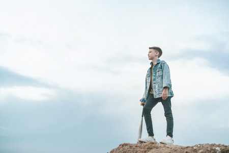 low angle view of boy in denim jacket standing with baseball bat and looking away at cloudy dayの写真素材
