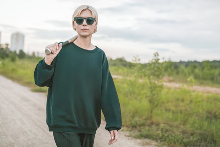 beautiful blonde woman in sunglasses holding baseball bat while walking on ground roadの写真素材