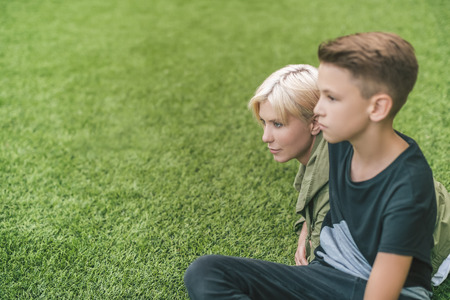 side view of beautiful mother and son looking away while sitting together on grassの写真素材
