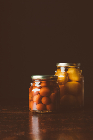 glass jars with preserved red and yellow tomatoes on wooden table in dark kitchenの写真素材