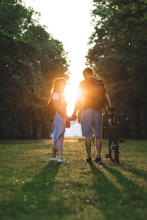 couple with backpacks and bicycle standing on meadow with setting sun behindの写真素材