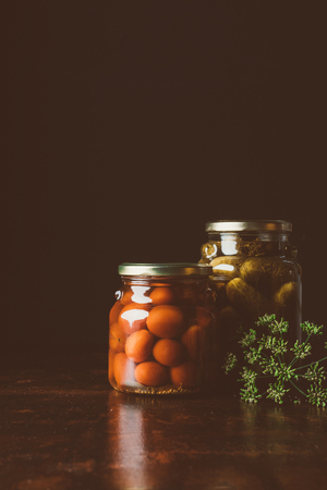 glass jars with preserved tomatoes and cucumbers on wooden table in dark kitchenの写真素材