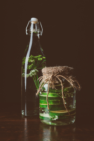 glass bottle with water and dill and glass jar with zucchini on wooden table on blackの写真素材