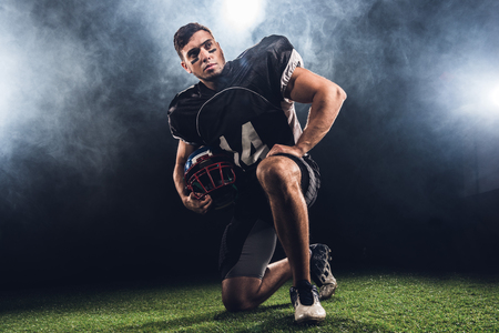 handsome young american football player with helmet standing on knee on blackの写真素材