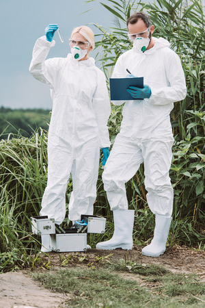 male scientist in protective suit and mask writing in clipboard while his female colleague examining sample of water in test flask outdoorsの写真素材
