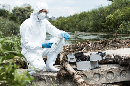 male scientist in protective suit holding sample of soil by tweezers and water in test flask...