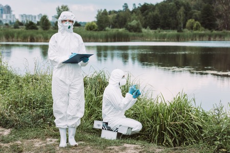 selective focus of female scientist in protective mask and suit writing in clipboard while her colleague putting sample of water near river outdoorsの写真素材