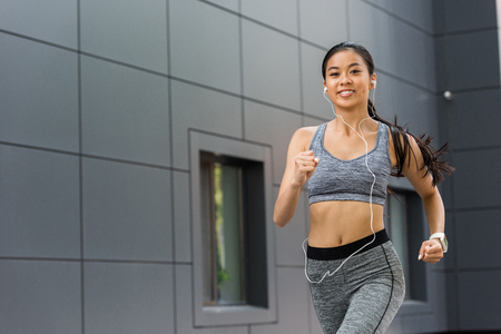 smiling young asian sportswoman in earphones running at city streetの写真素材