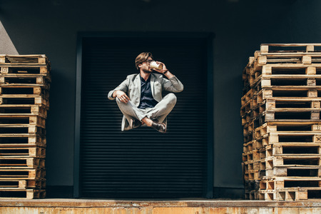 handsome young businessman floating in air in lotus pose and drinking coffee at industrial location...