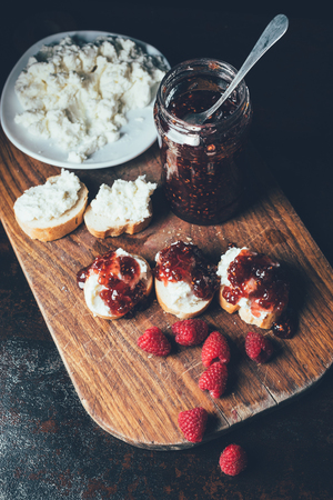 high angle view of raspberries, jar with jam, sandwiches and cream cheese on plate on cutting board