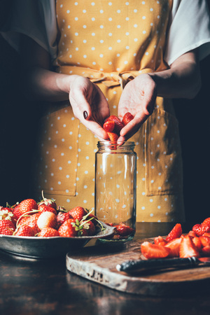 partial view of woman in apron putting strawberries in jar for cooking jamの写真素材