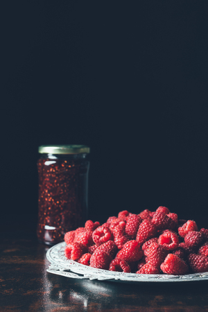 close up view of pile of raspberries in silver tray and jam in jar on blackの写真素材