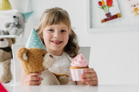 smiling birthday kid showing cupcake and holding teddy bear in cone at tableの写真素材