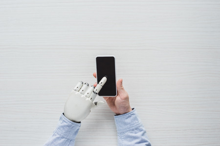 cropped image of man with cyborg hand using smartphone with blank screen over wooden table
