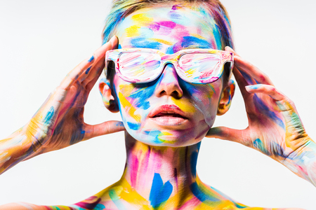 attractive girl with colorful bright body art and sunglasses touching head isolated on whiteの写真素材