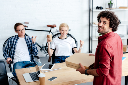 smiling young colleagues looking at happy man holding pizza boxes in officeの写真素材