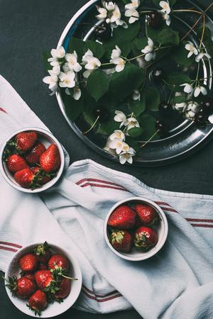top view of ripe red strawberries in bowls, towel and jasmine flowers on blackの写真素材
