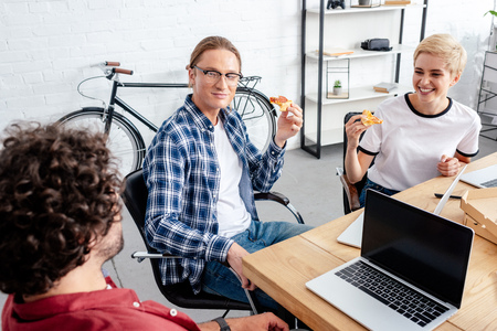 high angle view of smiling young coworkers eating pizza together while working together in officeの写真素材