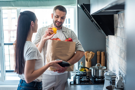married couple with paper package full of fresh vegetables for dinner in kitchen at homeの写真素材