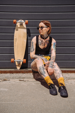 stylish woman with tattoos sitting near skateboard at streetの写真素材