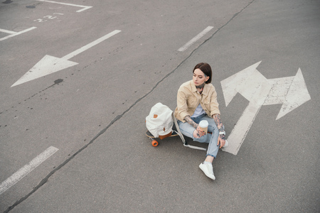 high angle view of tattooed woman sitting on skateboard with coffee cup at parking lotの写真素材