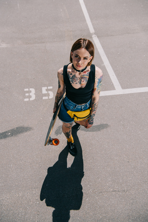 high angle view of stylish tattooed girl holding skateboard and looking at camera at parking lotの写真素材