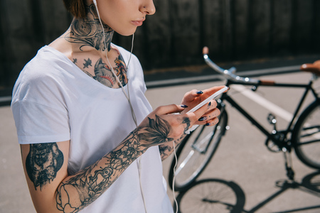 cropped image of tattooed woman in earphones listening music with smartphone near bicycleの写真素材