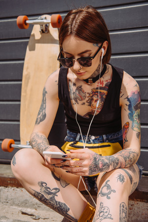 young tattooed woman in earphones listening music with smartphone near skateboardの写真素材